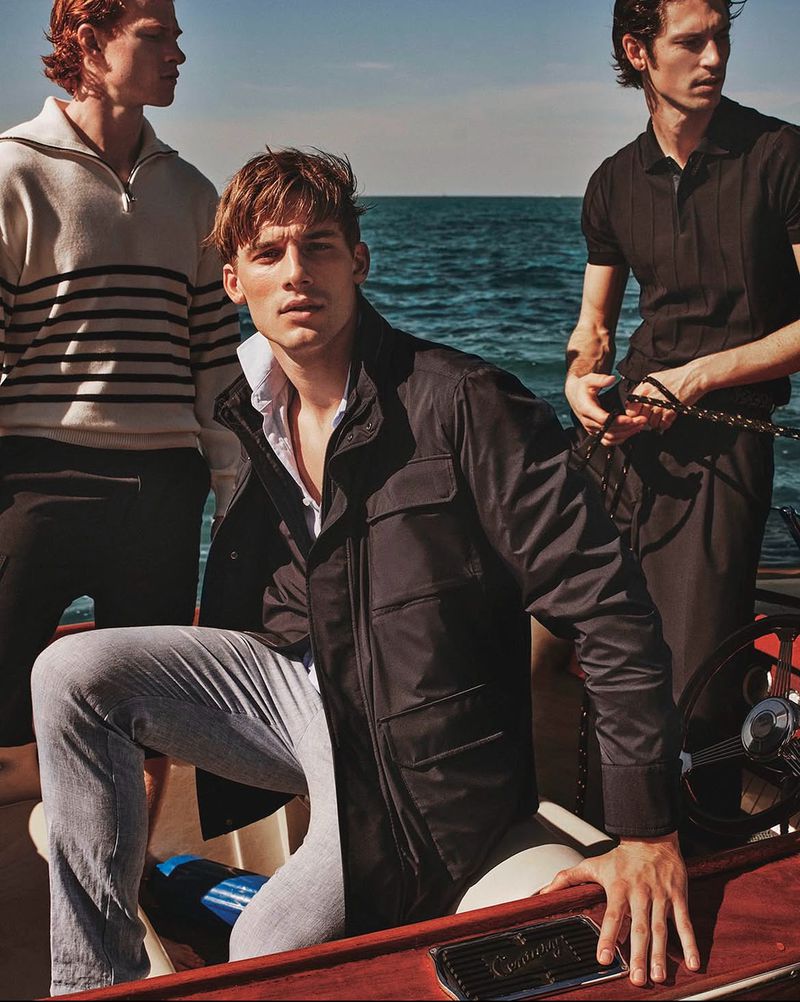 Three fair-skinned men in fashionable, nautical-themed clothing pose on a boat against an ocean backdrop. Clothing includes a zip-up sweater and jackets.