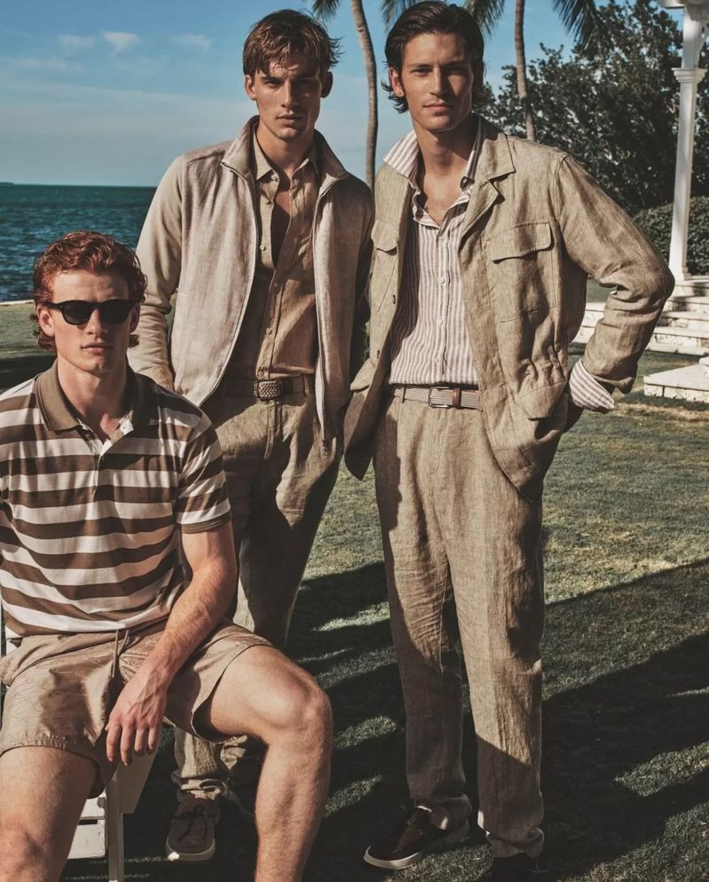 Three male fashion models pose outdoors near the sea. Two stand in linen suits, one sits wearing a striped shirt and shorts.