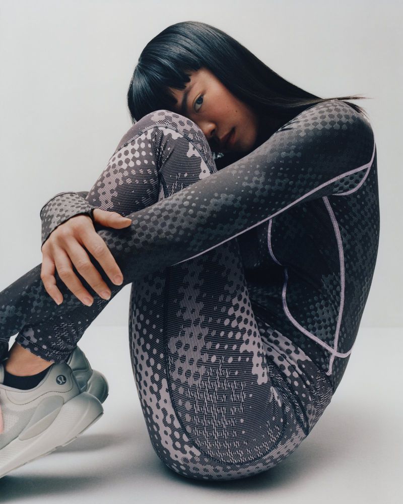 Woman in patterned athletic wear sits in studio. She is Asian, with black hair and white running shoes. The image features studio lighting.