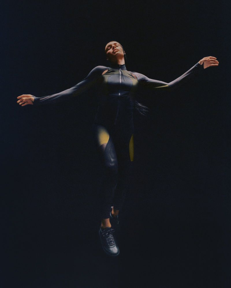 Athletic woman in black and yellow bodysuit and sneakers jumping in a dark studio with outstretched arms. Sport and fashion concept.