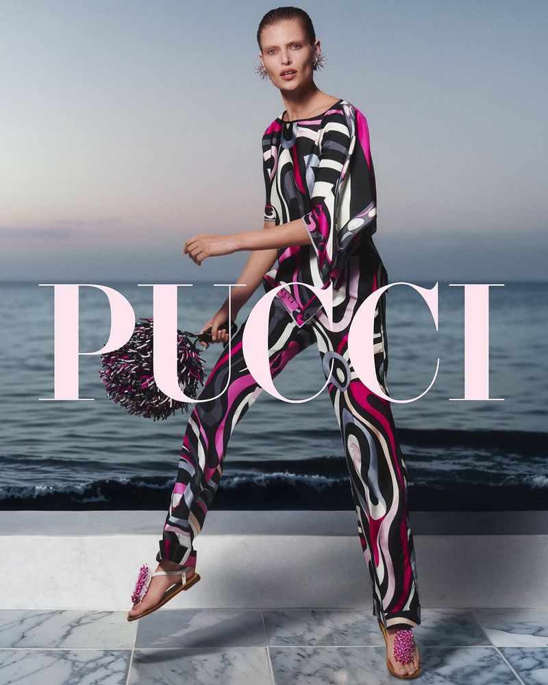 A woman in a patterned jumpsuit, holding a pom-pom bag, stands on a marble surface with the ocean and sky in the background.
