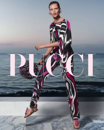 A woman in a patterned jumpsuit, holding a pom-pom bag, stands on a marble surface with the ocean and sky in the background.