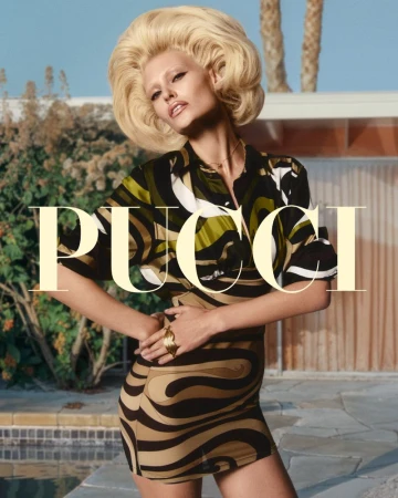 Fashion editorial shot of a fair-skinned woman with a blonde bouffant wearing a patterned outfit, set against an outdoor background.