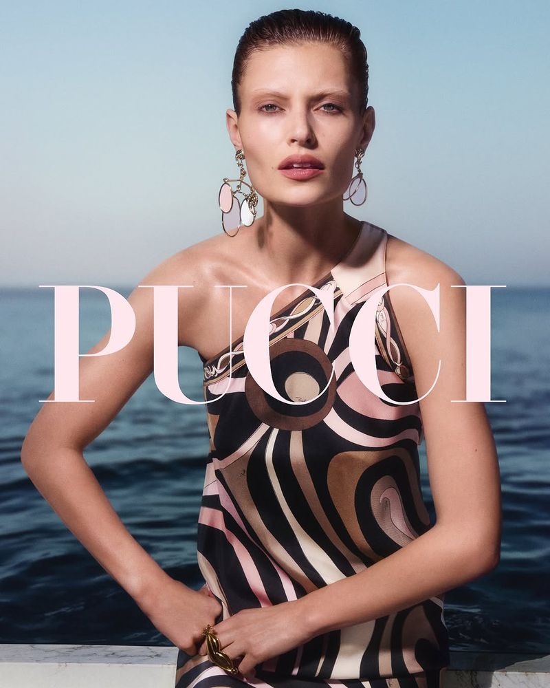 A model poses wearing a patterned dress in front of a seascape with the brand PUCCI overlaid in pink text, creating a fashion editorial shot.