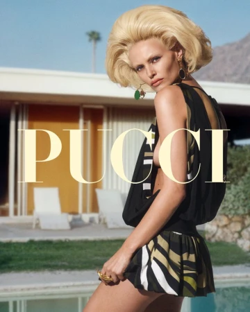 Fashion editorial image of a blond adult female model wearing a colorful printed mini dress in front of a modern house with a mountain backdrop.
