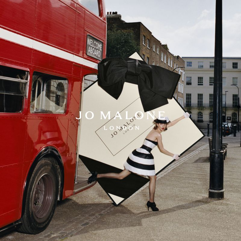 Fashion model in London street with red bus and oversized perfume box. Jo Malone ad. Stripes, bows, buildings, and outdoor ambiance.