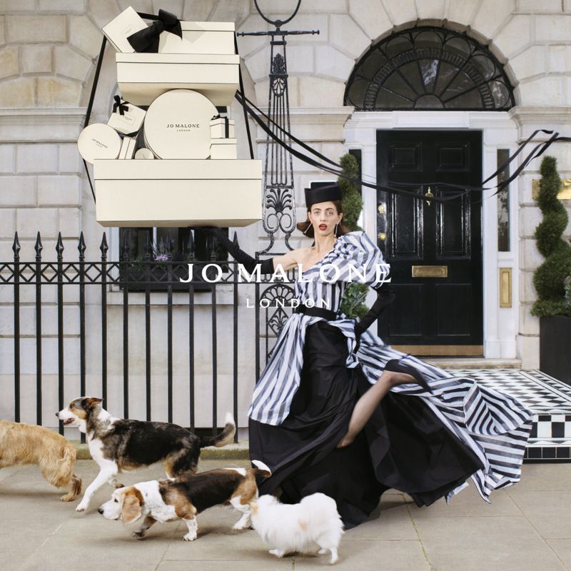 Fashion model in a black and white dress poses with Jo Malone boxes, surrounded by dogs on a city street with a building backdrop.
