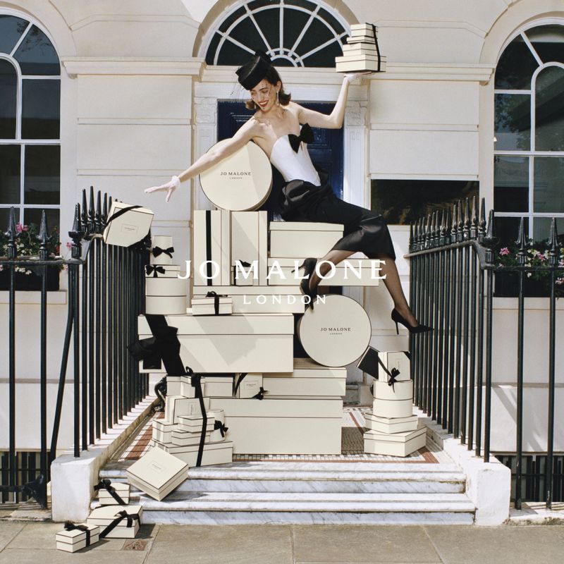 Elegant woman in black and white couture with hat strikes a pose among stacked Jo Malone gift boxes on a townhouse front staircase.