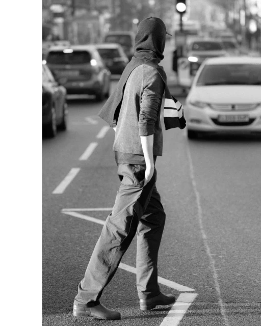 Monochrome image of a person in dark clothing crossing an urban street, with several cars in the background and surrounding buildings.