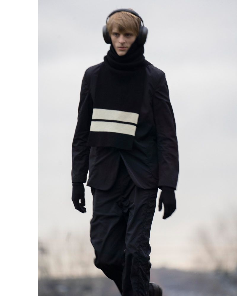 Man walking outdoors wearing headphones, black jacket, black pants, and a black scarf with white stripes for fashion concept.