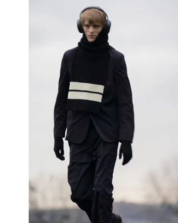 Man walking outdoors wearing headphones, black jacket, black pants, and a black scarf with white stripes for fashion concept.
