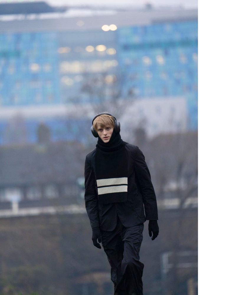 Model wearing black jacket and striped scarf outdoors against a blurred industrial background