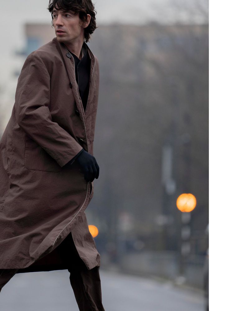 A person with dark hair walks outside wearing a long brown coat and gloves against a blurred urban background with light poles.