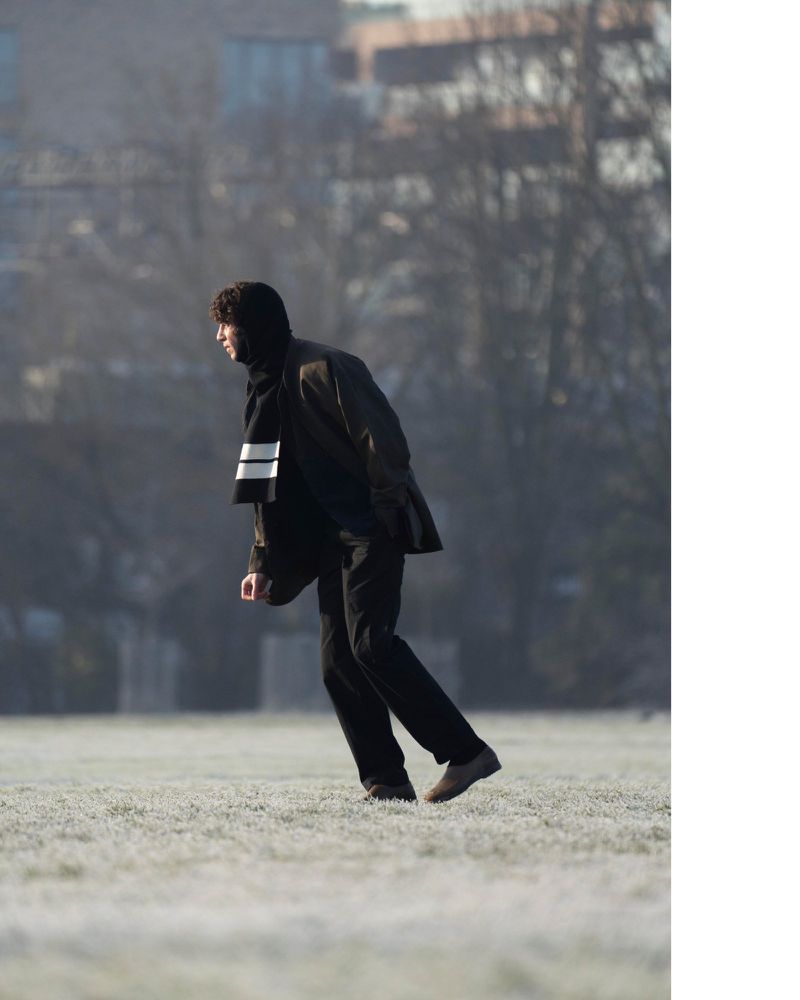 Adult male in jacket and striped scarf walks in a winter park with frosted grass, bare trees, and city buildings in soft-focus background.