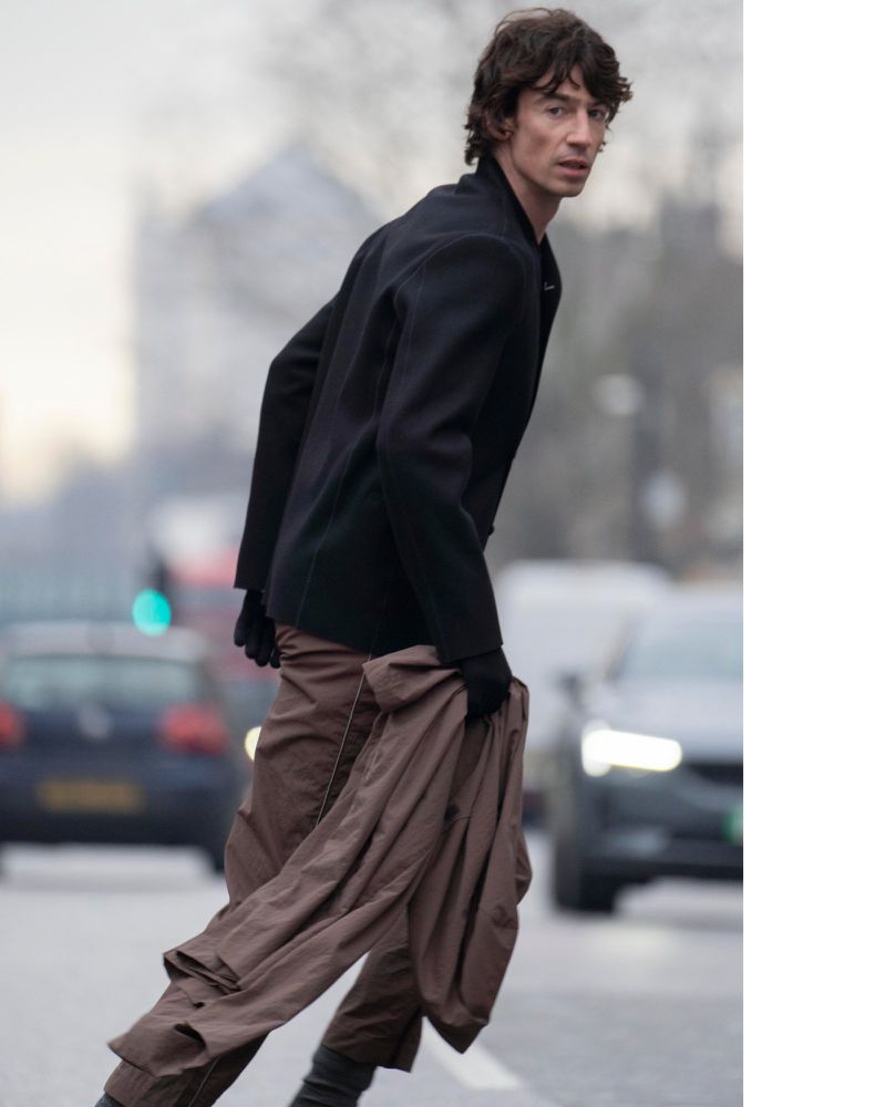 Man in black jacket and brown pants crosses street in city. Cars in background. Medium shot, eye-level angle, daytime, outdoor.
