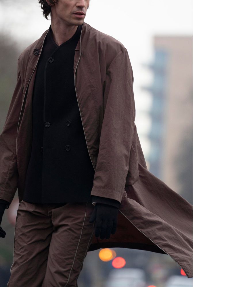 A fashionable adult male walks on a city street wearing a brown jacket and pants with a black buttoned jacket and black gloves.
