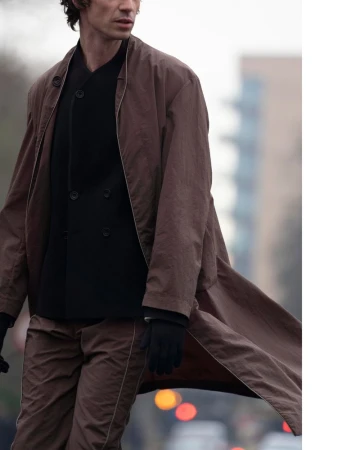A fashionable adult male walks on a city street wearing a brown jacket and pants with a black buttoned jacket and black gloves.
