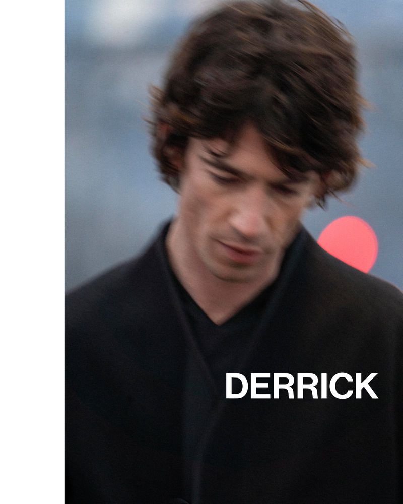 Close-up portrait of a man wearing a dark V-neck jacket with Derrick branding text against a blurred outdoor background.