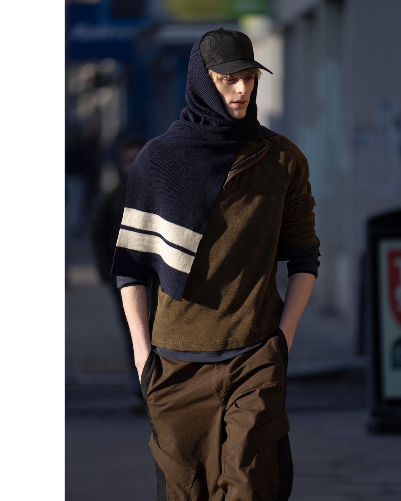 Model wearing a dark cap, hooded scarf, and brown layered outfit walking outdoors for Derrick Fall/Winter 25.