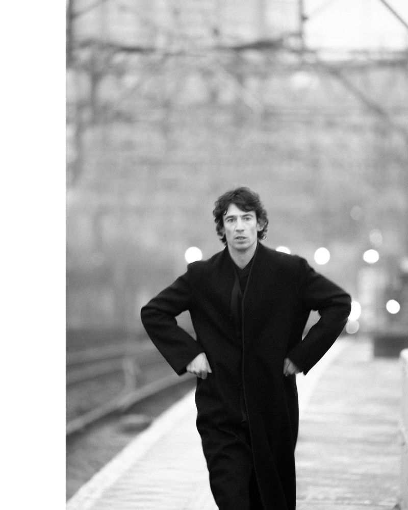 Monochrome portrait of a man in a long coat standing on a train platform with hands on his hips, waiting with blurry background details.