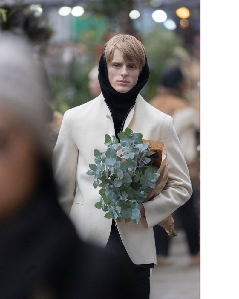 Man in off-white jacket holds wrapped bouquet in outdoor setting with soft light and blurred background.