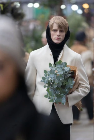 Man in off-white jacket holds wrapped bouquet in outdoor setting with soft light and blurred background.
