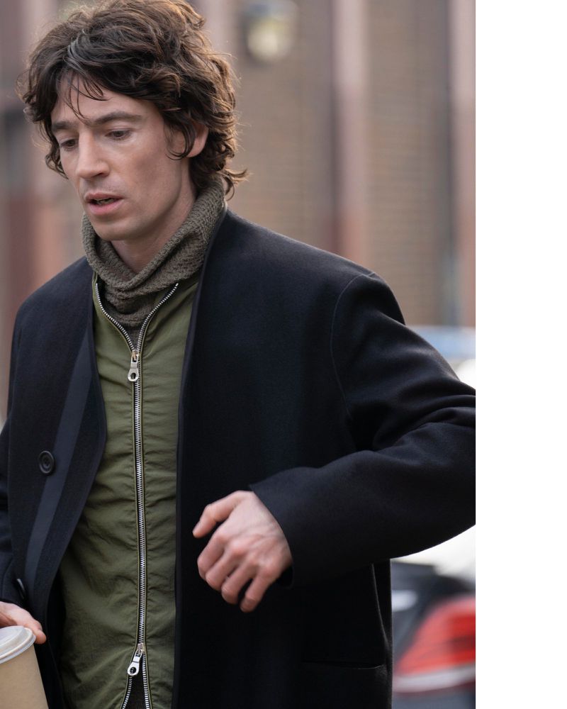 Adult male with wavy brown hair in a black overcoat and green jacket outdoors on a city street holding a disposable coffee cup.