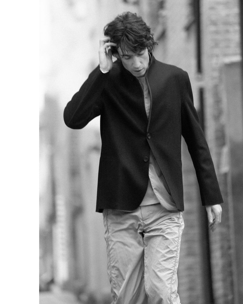 Monochrome portrait of a man walking outdoors wearing a dark blazer, light trousers, and adjusting his hair.