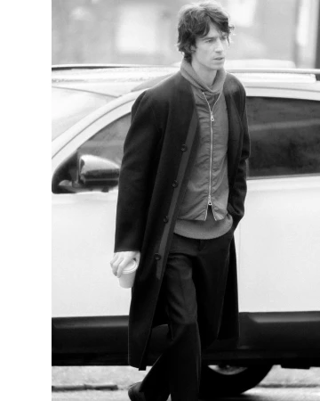 Monochrome image of a male model walking in front of a car wearing a long dark coat and zipped jacket