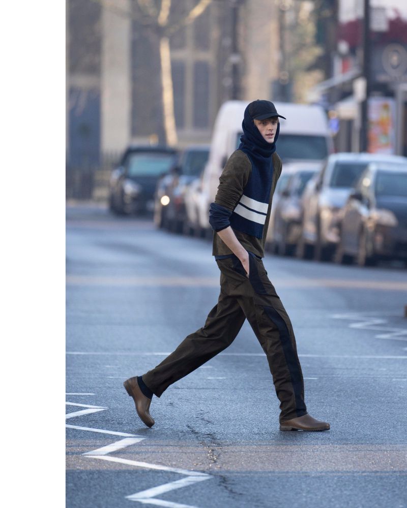 Man in a dark jumpsuit and hat crossing a street with cars in the background, captured in a medium shot on a city street.
