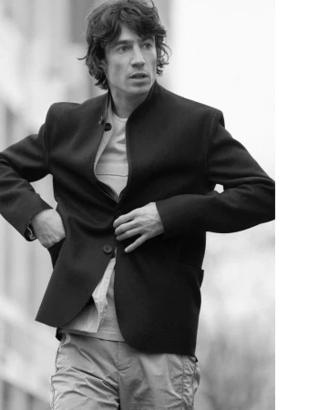 Black and white portrait of a man wearing a dark jacket over a light shirt against an out-of-focus urban background.