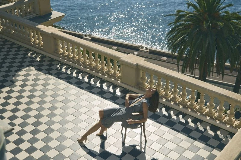 Woman in a grey dress reclines on a chair on a checkered balcony, overlooking the ocean and palm trees in bright daylight.