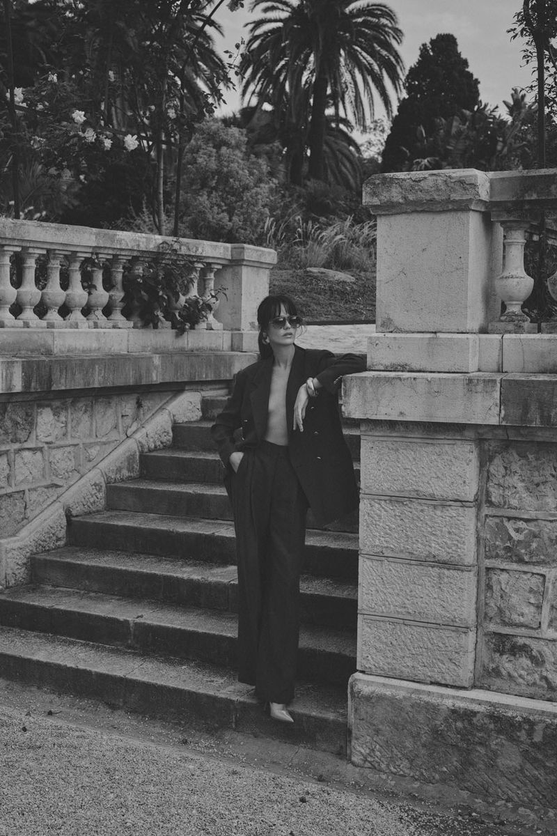 A woman in a dark pantsuit poses on stone stairs in front of palm trees, with a balustrade and greenery in a monochrome outdoor setting.