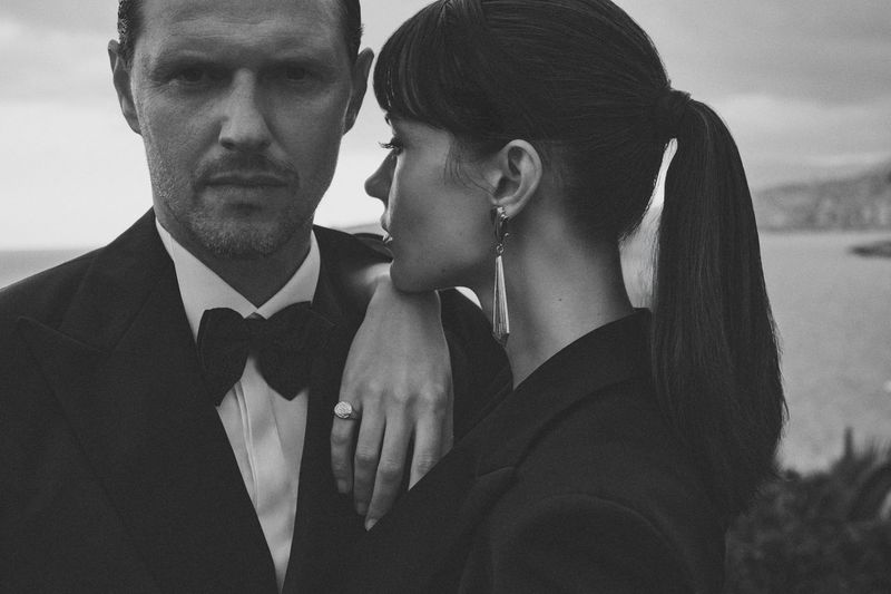 Monochrome portrait of a stylish couple outdoors: A man in a tuxedo and a woman with long earrings and a ponytail strike a fashionable pose.