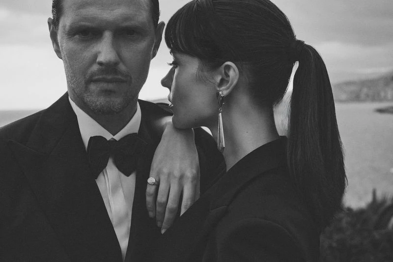 Monochrome portrait of a stylish couple outdoors: A man in a tuxedo and a woman with long earrings and a ponytail strike a fashionable pose.