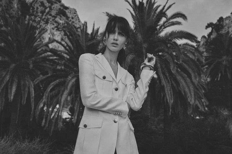 Monochrome fashion portrait of a female model wearing a white utility jacket outdoors against palm trees.