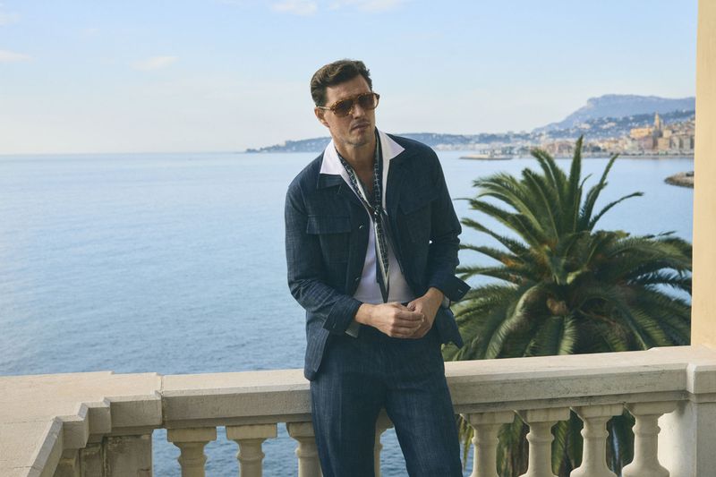 Man in a blue jacket and pants stands on a stone balcony overlooking the ocean and a coastal city with palm trees on a sunny day.