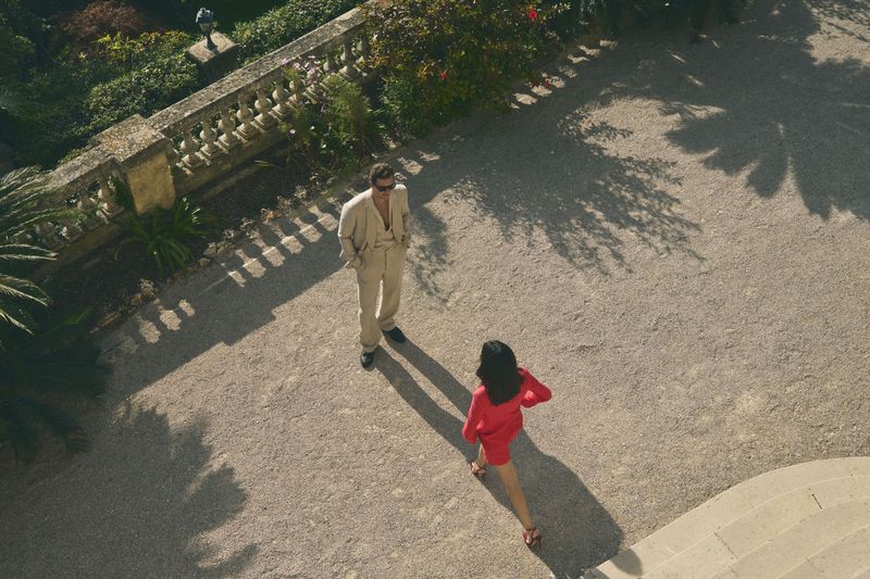 Aerial view of an adult man in a beige suit and sunglasses standing near an adult woman in a red dress in a sunny outdoor courtyard.