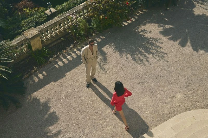 Aerial view of an adult man in a beige suit and sunglasses standing near an adult woman in a red dress in a sunny outdoor courtyard.