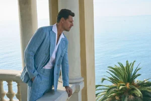 Man wearing a light blue striped suit leaning on a balcony overlooking the sea