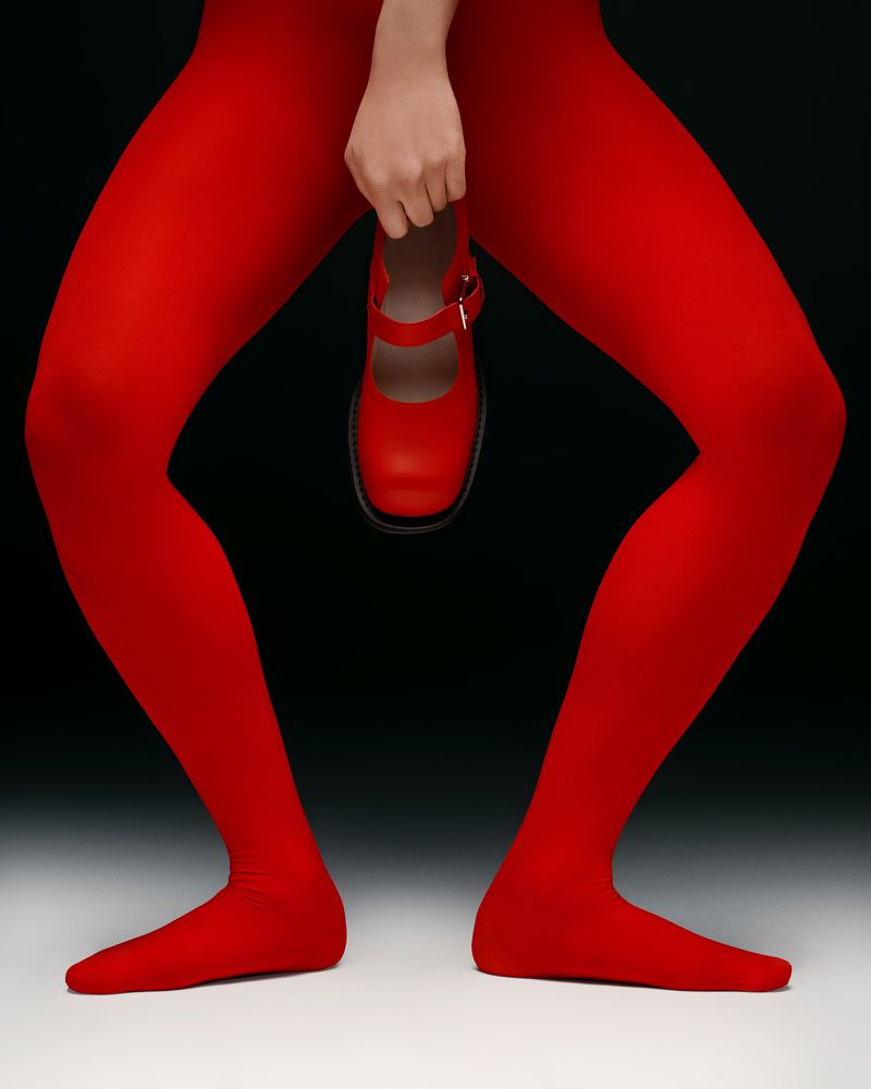 A posed studio shot featuring a person's legs wearing red tights, holding a red Mary Jane style shoe against a dark studio background.