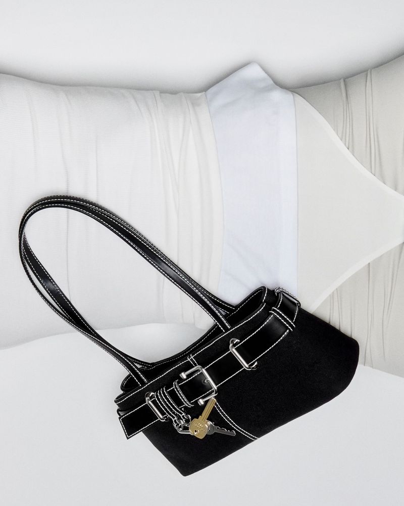 Studio shot of a black purse with silver hardware and keys, resting on white pillows with soft, neutral-toned bedding in background.
