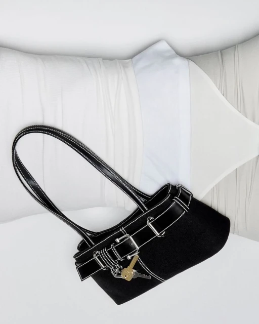 Studio shot of a black purse with silver hardware and keys, resting on white pillows with soft, neutral-toned bedding in background.