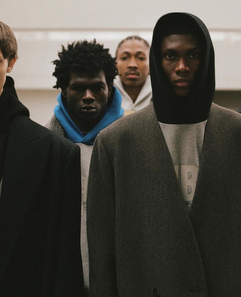 Men in coats and hoodies stand in a line indoors, creating a modern fashion editorial showcasing urban style and diverse representation.
