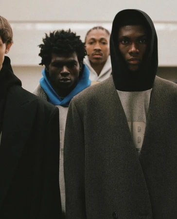 Men in coats and hoodies stand in a line indoors, creating a modern fashion editorial showcasing urban style and diverse representation.