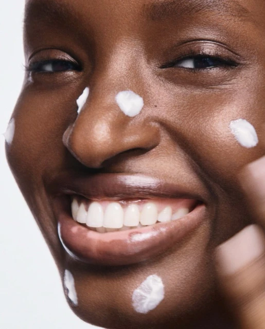 Close-up of a smiling African American woman's face with skincare product dots on her face against a bright white background.