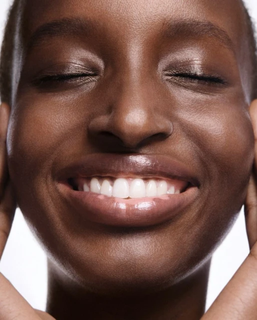 Close-up shot of a dark-skinned woman with her eyes closed and a happy expression, showcasing her teeth and skin texture.