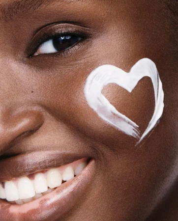 Close-up of a smiling Black woman with flawless skin, adorned with a heart shape drawn on her cheek with white moisturizing cream.