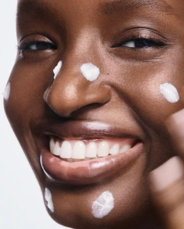 Close-up of a smiling black woman with dots of white moisturiser cream on her face against a plain background