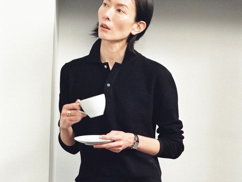 Woman in a black shirt holds a white cup and saucer in a bright, minimalist interior, showcasing a blend of fashion and lifestyle.